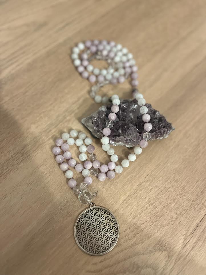 collier mala en pierre naturelles de kunzite de pierre de lune et de cristal de roche avec son pendentif fleur de vie posé sur une druze d'améthyste