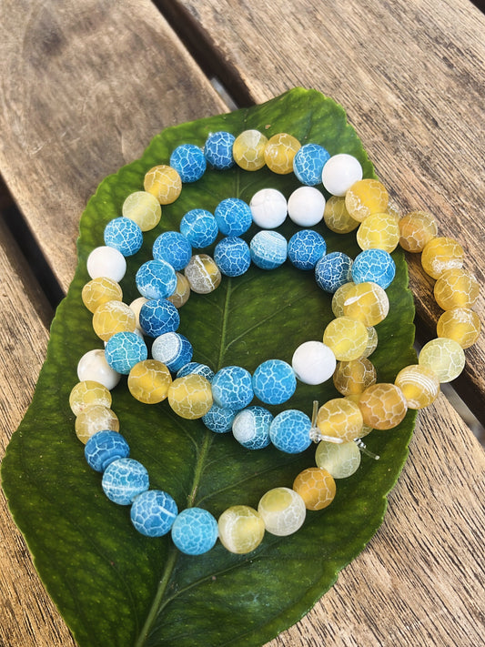 trois bracelets en perles jaune, bleu et pierres naturelles de howlite posés sur une feuille verte sur une table en bois.